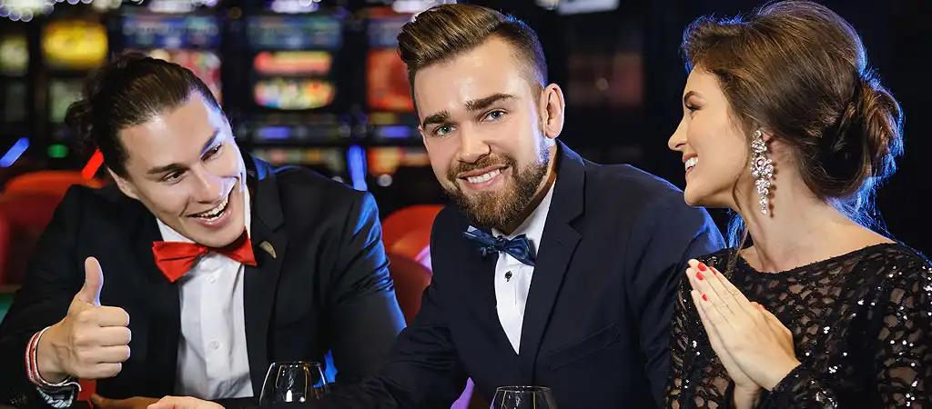 Három elegánsan öltözött játékos beszélget és nevet egy kaszinóban, élvezve a társaságot és a játékot.